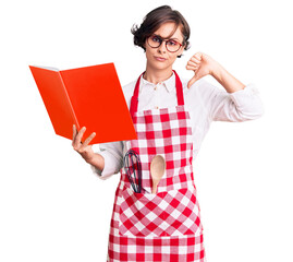 Wall Mural - Beautiful young woman with short hair wearing professional baker apron reading cooking recipe book with angry face, negative sign showing dislike with thumbs down, rejection concept