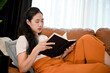 © bongkarn - Peaceful young Asian female leans on sofa while reading a book in the living room.
