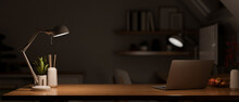 Modern Dark Wooden Office Desk At Night Under The Warm Light From Table Lamp With Laptop