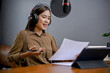Talented Asian female freelance blogger or radio host reading her podcast script in her studio.
