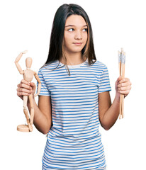 Canvas Print - Young brunette girl with long hair holding small wooden manikin and pencils smiling looking to the side and staring away thinking.