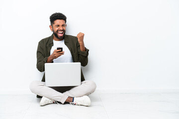 Wall Mural - Young Brazilian man with a laptop sitting on the floor isolated on white with phone in victory position