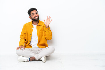 Young Brazilian man sitting on the floor isolated on white background saluting with hand with happy expression