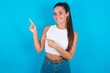 beautiful brunette woman wearing white tank top over blue background indicating finger empty space showing best low prices, looking at the camera