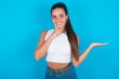 © Roquillo - Funny beautiful brunette woman wearing white tank top over blue background hold open palm new product great proposition