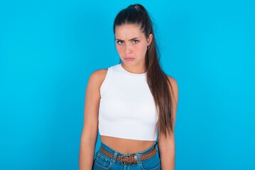 Wall Mural - Displeased upset beautiful brunette woman wearing white tank top over blue background frowns face as going to cry, being discontent and unhappy as can't achieve goals,  Disappointed model has troubles