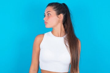 Close up side profile photo beautiful brunette woman wearing white tank top over blue background not smiling attentive listen concentrated