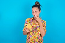 Beautiful Brunette Woman Wearing Colourful Shirt Over Blue Background Being Deeply Surprised, Stares At Smartphone Display, Reads Shocking News On Website, Omg, Its Horrible!