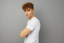 A Calm, Curly-haired Young Man Stands In A White T-shirt On An Isolated Background Sideways To The Camera, Facing The Viewer And Looking With Folded Arms On His Chest