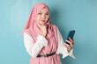© Reezky - Portrait of a thoughtful young Muslim woman wearing pink hijab looking aside while holding smartphone