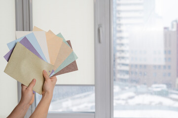 Wall Mural - Woman holding fabric samples of roller blinds against window background. 