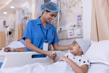 Young nurse visiting and consoling little girl lying on bed. Female child patient is looking at care giver while resting in ward. They are at healthcare center.
