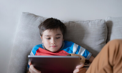 Wall Mural - School kid using tablet readying story from internet, Happy boy sitting on sofa doing studying online learning  at home, Child holding digital pad using wifi playing game or chatting with friends.