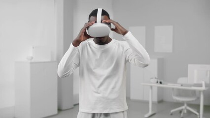 Poster - Metaverse. concept. Portrait of African-American man in white clothes put on augmented reality headset standing in room with white walls and furniture. 