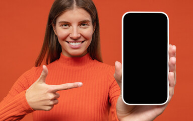 Portrait of happy young woman pointing to blank phone screen, isolated on orange background