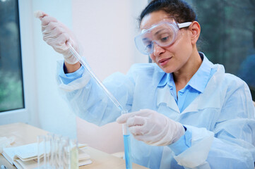 Latin American pharmacologist, clinical researcher using a graduated glass pipette, drips a chemical substance into a test tube in science laboratory. Scientific investigation in medical field