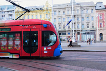 Closeup Of Red And Blue Tram In The Popular Jelacic Square In Downtown Zagreb. Old Architectural Background. Tourism And Travel To Croatia Concept. Soft Blurred Urban Background With Tourists
