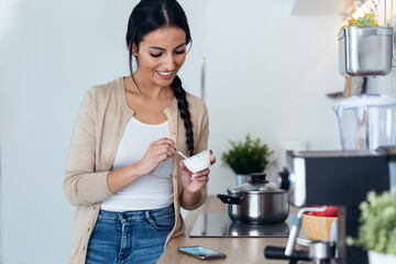 Pretty woman sending messages with mobile phone while eating yogurt int he kitchen at home.