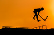 Unrecognizable teenage boy silhouette showing high jump tricks on scooter against orange sunset sky at skatepark. Sport, extreme, youth, urban culture, freestyle, outdoor activity concept