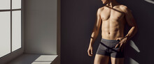 Attractive Young Man In Underwear Standing Near Window. Shirtless Male Model With Fit Body Posing In Underpants. Banner, Header Background. Cropped Shot In Bright Sun Light With Contrasting Shadows