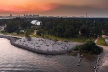 Russia, St. Petersburg, 17 August 2022: Observation Deck On The Embankment Of Krestovsky Island At Sunset, People Have Fun In Summer Evenings, Reflection Of The Sky On The Water, Benches For Rest