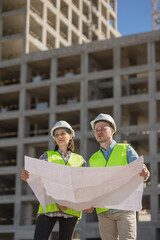 Wall Mural - two engineers in protective vests and white helmets on the background of a house under construction look at the object's circuit board or diagram. European male and female engineers perform fire