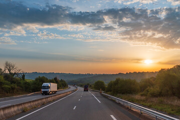 Wall Mural - Sunrise on a highway surrounded by trees with several trucks circulating.