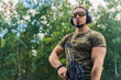 © PoppyPix - Medium outdoor shot of a military-looking caucasian muscular man in moro t-shirt looking far away at his target and holding black rifle with a loupe to look through. High quality photo