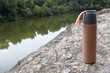Beige thermos bottle and cup of tea or coffee on rock over the river. Beautiful summer, spring, autumn landscape. Picnic on the rock