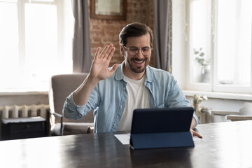 Wall Mural - Happy millennial freelance business man in casual talking to client on video call, waving greeting hand, saying hello, using tablet computer at work table, getting good news, smiling, laughing