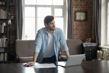 Serious Thoughtful Millennial Business Man Standing At Laptop And Documents, Leaning On Work Table In Home Office Loft Interior, Looking Away, Thinking Over Creative Ideas For Project, Making Decision