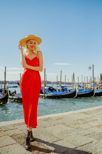 Happy Smiling Woman Wearing Straw Hat, Red Midi Dress Posing  On The Pier Of Grand Canal With Gondolas In Venice, Italy. Fashion, Travel, Lifestyle Conception. Full-length Outdoor Portrait