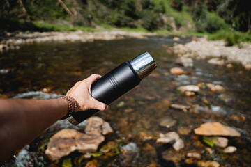 Wall Mural - The guy holds a thermos in his hand against the background of the river, his hand holds a thermo bottle. trekking on a mountain river alone, hiking in extreme conditions.