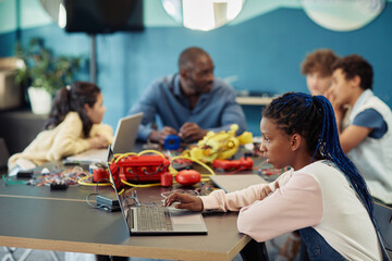 Wall Mural - Side view portrait of young black girl using laptop in engineering class and programming robots, copy space