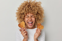 Horizontal Shot Of Happy Optimistic Woman Exclaims Loudly Holds Two Heart Shaped Waffles Has Unhealthy Nutrition Dressed In Casual Jumper Isolated Over White Background. Sweet Tasty Dessert.