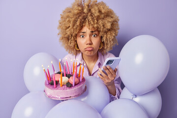 Canvas Print - Upset worried woman looks sadly at camera poses with festive cake and smartphone celebrates birthday surrounded by inflated balloons isolated over purple background. Festive occasion concept