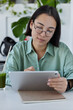 © wayhome.studio  - Smart Asian female student makes online project via touchpad connected to wireless internet searches useful information wears round eyeglasses and casual shirt poses indoor against cozy interior.