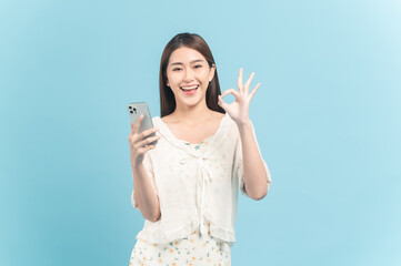 Wall Mural - Beautiful young asian woman in white dress with flower pattern using smartphone show okay sign isolated on blue background