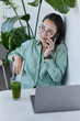 © wayhome.studio  - Asian woman works online has phone conversation with client works on modern laptop computer drinks fresh healthy smoothie poses in home office wears spectacles and shirt looks thoughtfully away