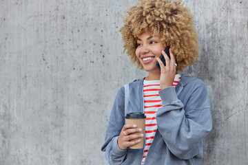 Beautiful curly haired teenage girl calls to friend during coffee break holds takeaway cup uses wireless internet for phoning wears hoodie poses against grey concrete wall looks happily away