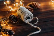 Close Up Of Rope Thread And Fir Pine Cone Decoration With With Christmas Tree Garland Lights Flickering In Background. Shallow Depth Of Field. Xmas Gift Card And Handmade Present Pack Concept.