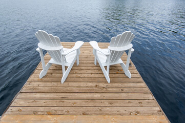 Two white Adirondack chairs on a cottage wooden dock facing the blue water of a lake in Muskoka, Ontario Canada.