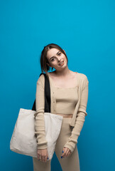 Wall Mural - Beautiful brunette woman in a beige costume with cotton bag on a shoulder on a blue background. Mockup and zero waste concept.