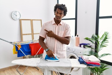 Sticker - African man with curly hair ironing clothes at home pointing with hand finger to the side showing advertisement, serious and calm face