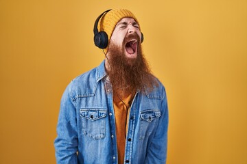 Canvas Print - Caucasian man with long beard listening to music using headphones angry and mad screaming frustrated and furious, shouting with anger. rage and aggressive concept.