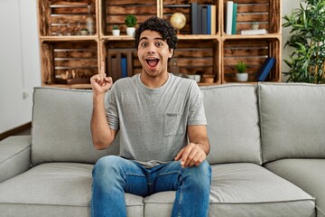 Sticker - Young hispanic man wearing casual clothes sitting on the sofa at home pointing finger up with successful idea. exited and happy. number one.