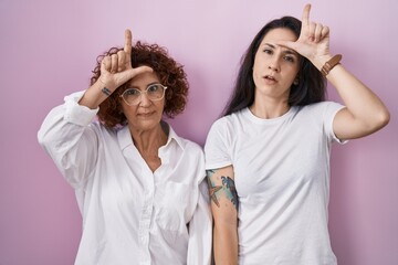 Hispanic mother and daughter wearing casual white t shirt over pink background making fun of people with fingers on forehead doing loser gesture mocking and insulting.