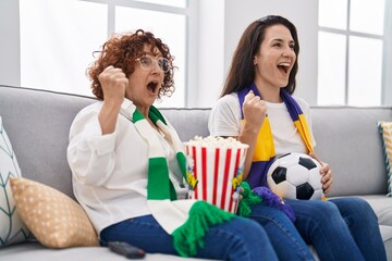 Poster - Hispanic mother and daughter watching football supporting team screaming proud, celebrating victory and success very excited with raised arms