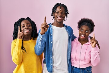 Sticker - Group of three young black people standing together over pink background pointing finger up with successful idea. exited and happy. number one.