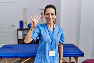 Canvas Print - Young hispanic woman wearing physiotherapist uniform standing at clinic showing and pointing up with fingers number four while smiling confident and happy.